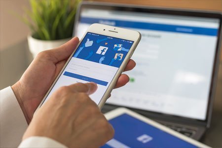 Person holding an iphone with Facebook on the screen over a laptop with Facebook homepage