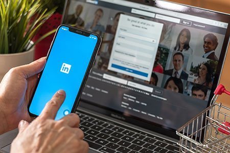 Finger pointing to phone with LinkedIn logo on screen over laptop with Linkedin on homepage, a small shopping cart and a plant on the table