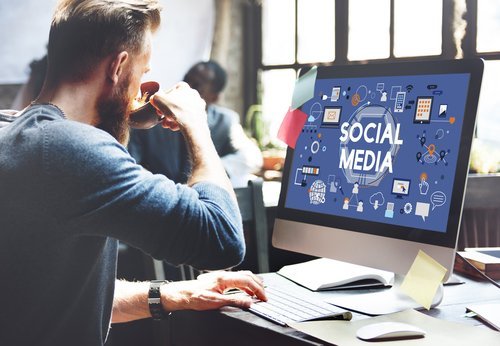 Male employee drinking coffee at office desk while working on social media strategy at desktop