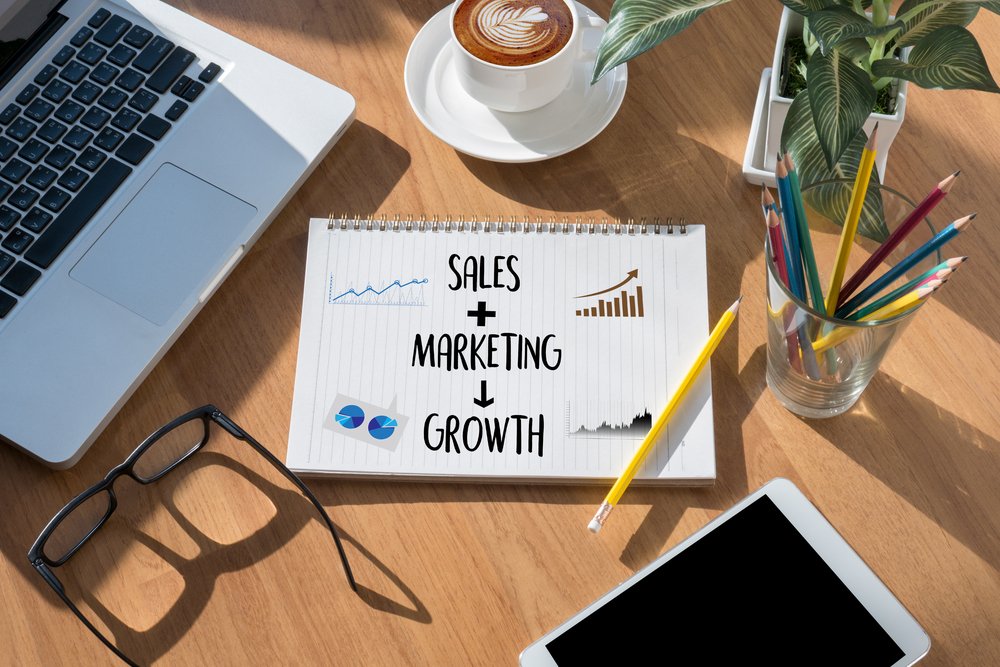 Desk littered with laptop keyboard, pencils, glasses, a coffee, and a notebook with the phrases sales, marketing, and growth written on it in black marker.